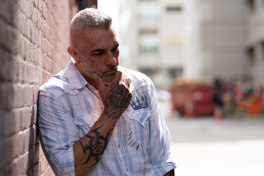 Tattooed middle-aged man leaning against a brick wall, looking thoughtful about sex and intimacy as part of men’s self improvement.
