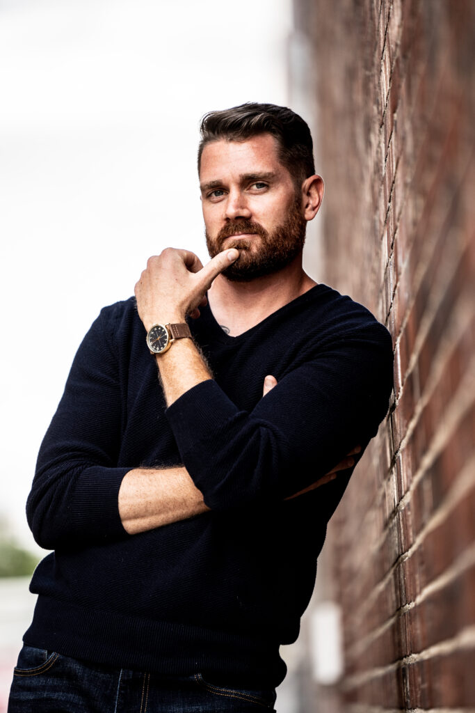 Bearded man in a dark shirt leaning against a wall, looking thoughtful about resilience, self repair, and men’s self improvement.