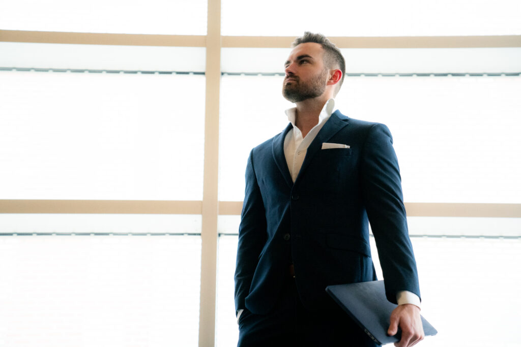 Confident man in a navy suit standing by a large window, symbolizing men’s self improvement and leadership.
